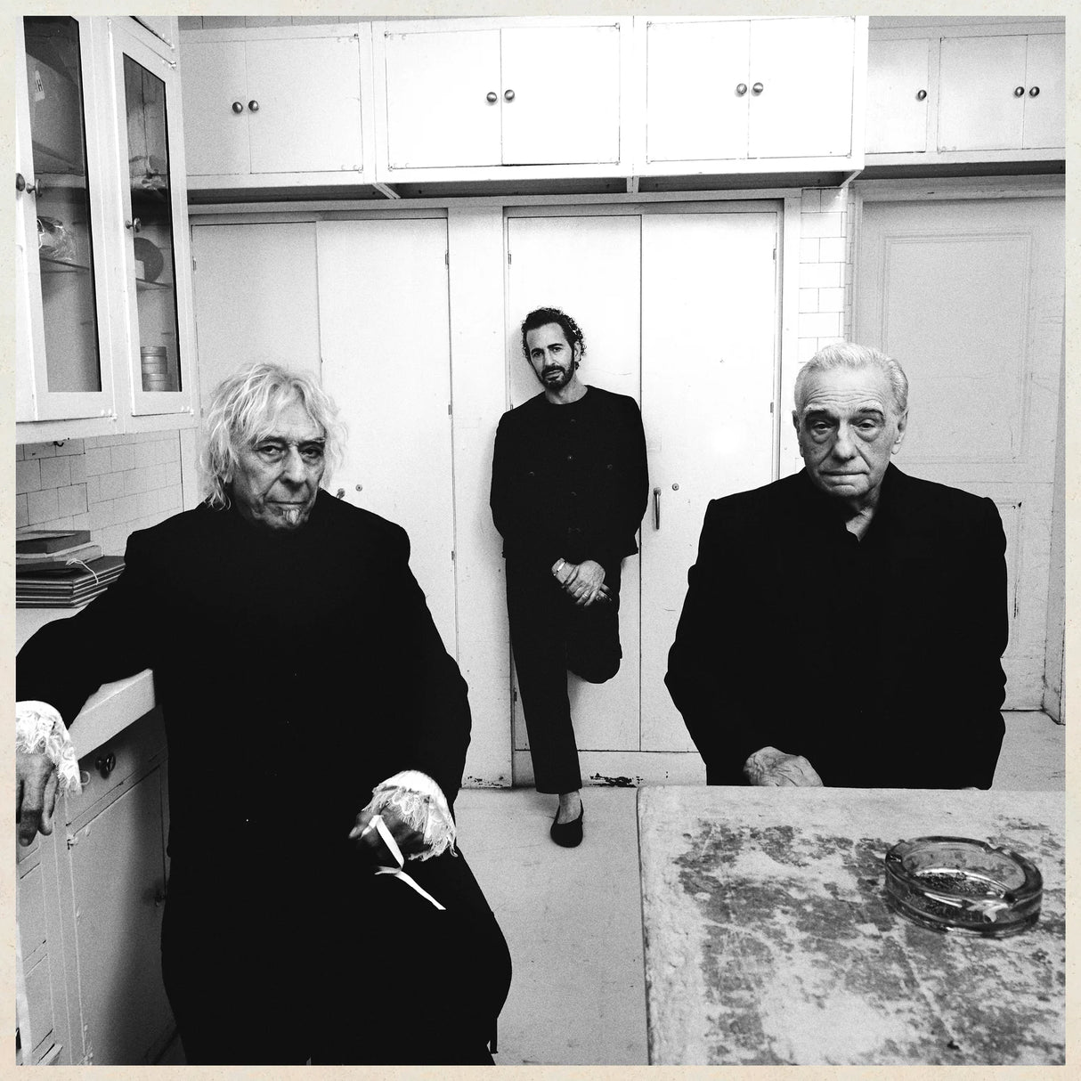Three people in a kitchen setting, with white cabinets and a tiled wall.