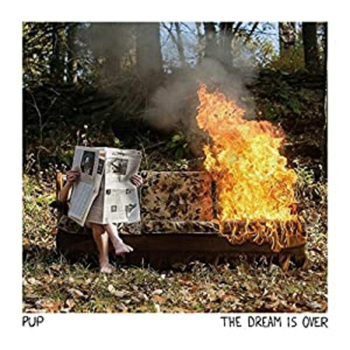 Person reading a newspaper next to a burning couch in a forest setting, album cover for 'The Dream is Over' by PUP.