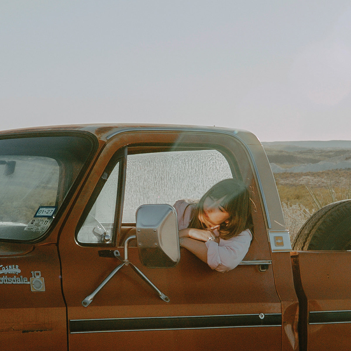 A woman with long brown hair rests her head on her arms inside the passenger window of a vintage rust-brown pickup truck, with a blurred outdoor landscape in the background.