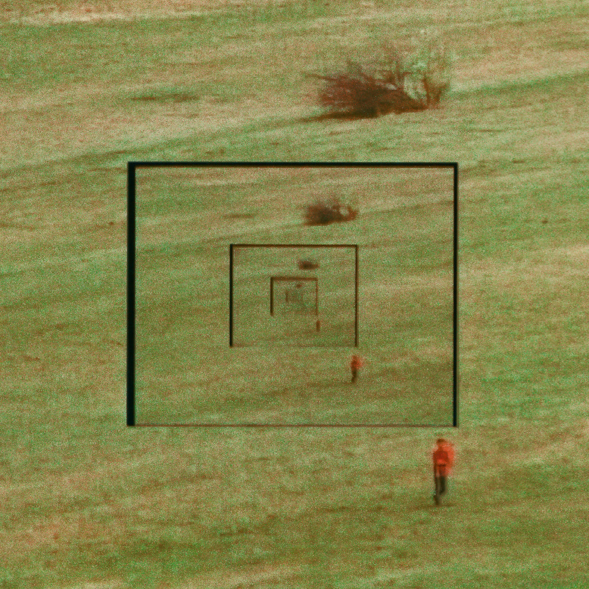 A square grassy field with concentric black target-like frames and a small person in an orange top standing near the bottom right, photographed from above.