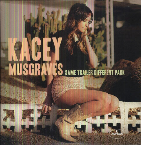 Album cover of Kacey Musgraves with the title 'Same Trailer, Different Park'. She is seated, wearing a black top and patterned shorts, with a desert landscape in the background.