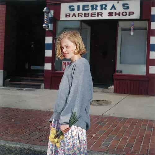 A person standing outside a barber shop with a sign that reads 'Sierra's Barber Shop'. They are holding a bouquet of flowers and wearing a grey sweater and a skirt with a floral pattern.