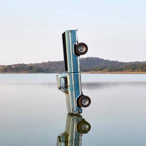 A silver-colored car, presumably the 'F-1 Trillion', partially submerged in water with a scenic view of a lake and mountains in the background.