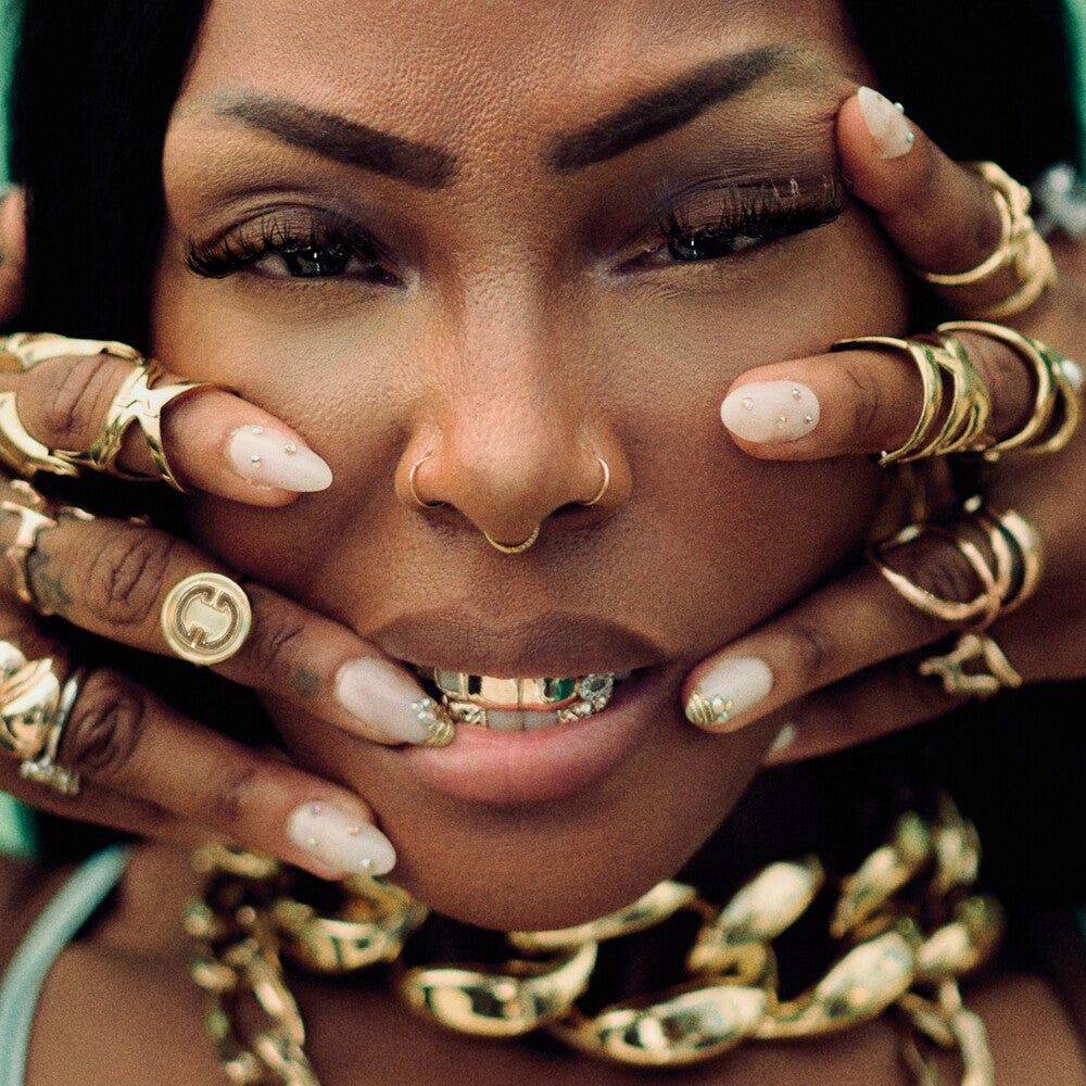 A close-up portrait of a woman with dark skin wearing an extensive collection of gold jewelry, including thick gold chain necklaces, multiple gold rings, and gold nose jewelry, with her hands framing her face.