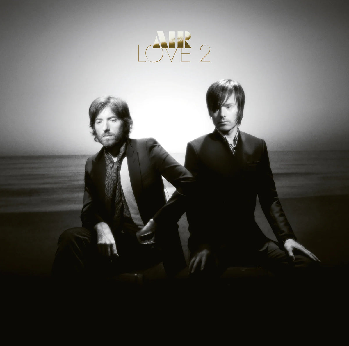 Album cover for Air's 'Love 2' featuring the band members seated on a dark surface against a bright, hazy sky background. The gold 'AIR' logo and 'LOVE 2' title are displayed at the top.