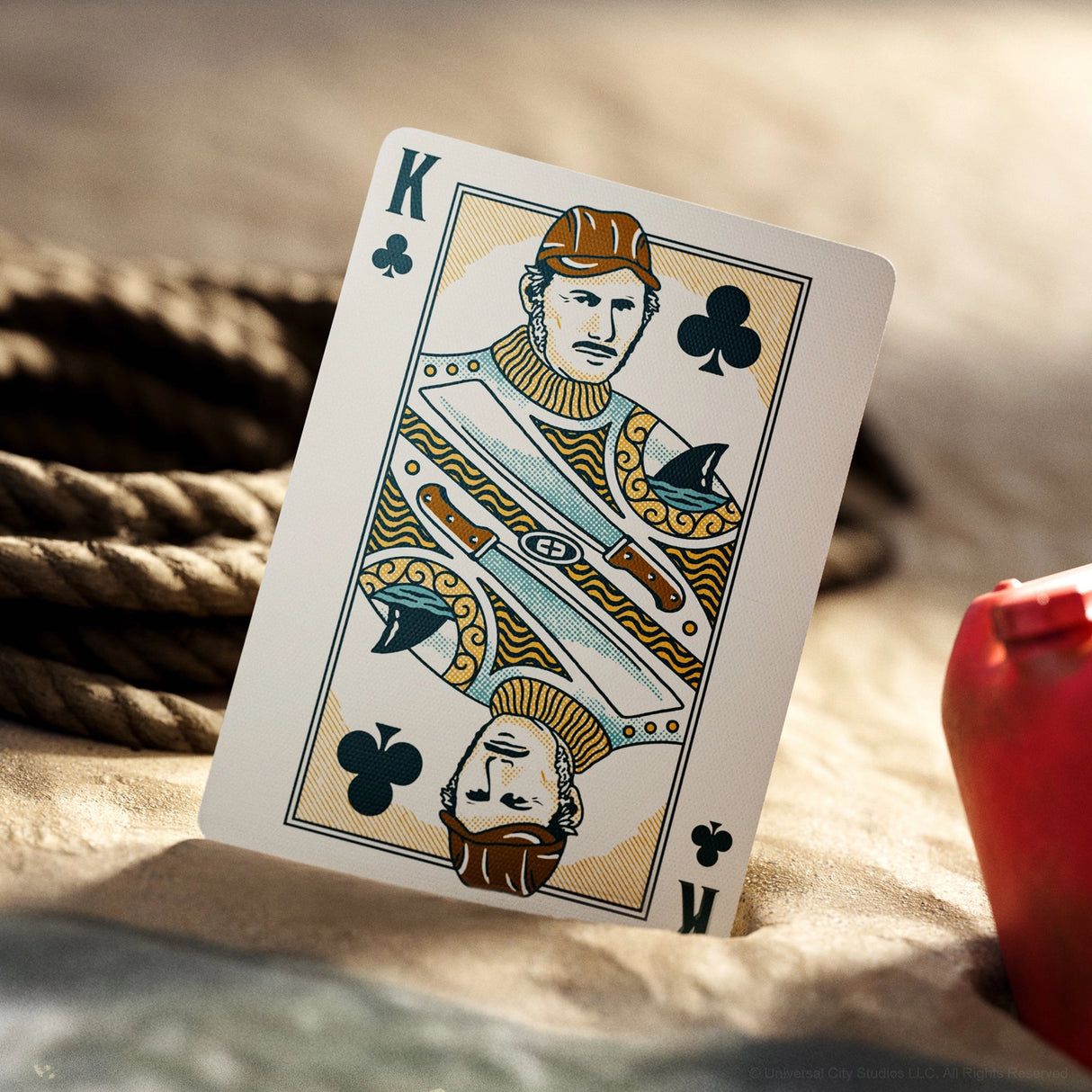 King of Clubs playing card on a sandy surface with ropes in the background