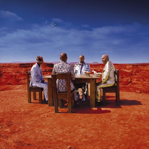 A group of four people seated around a table in an arid landscape with red soil and blue sky.