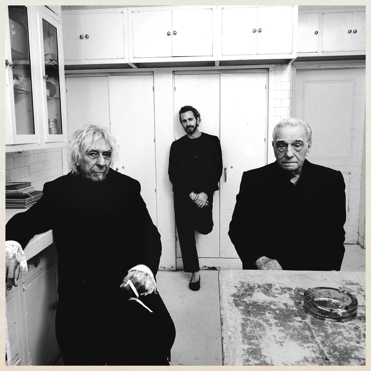 Three people in a kitchen setting, with white cabinets and a tiled wall.