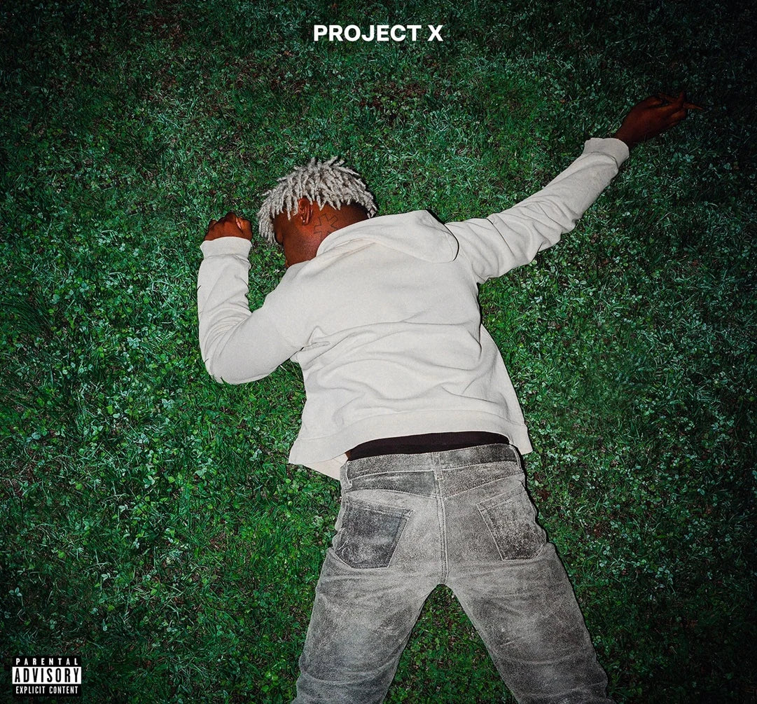 A person lying on green grass wearing a white long-sleeve shirt and faded gray jeans, with the text 'PROJECT X' at the top and a Parental Advisory label in the lower left corner.