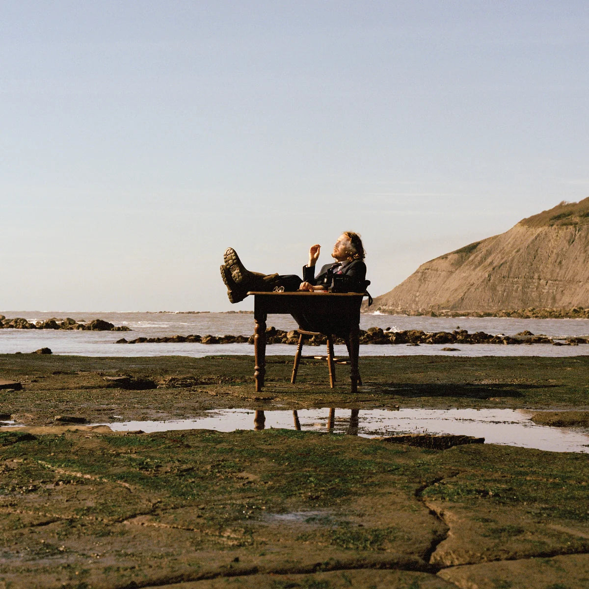 A person reclines on a small wooden table outdoors on a rocky shoreline with water, a pale sky, and a grassy cliff in the background.