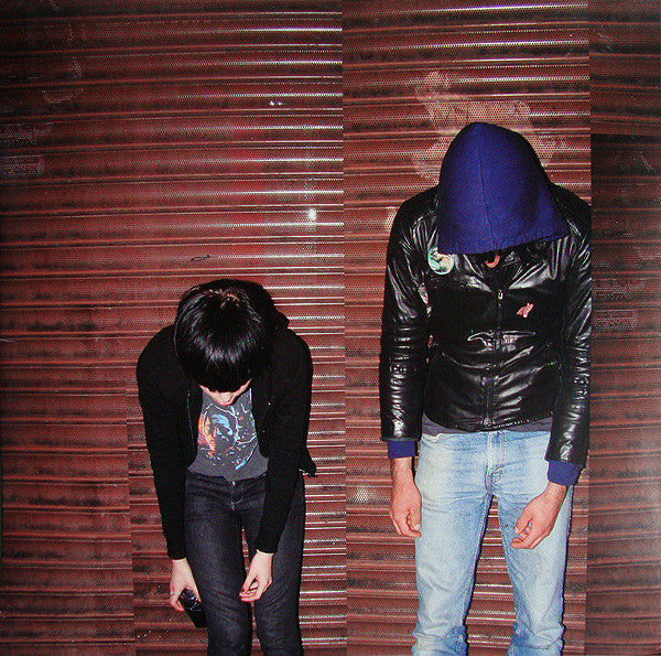 Two individuals, presumably members of the band Crystal Castles, standing in front of a corrugated metal wall. One is bending over while the other stands with a hoodie on, facing the camera.