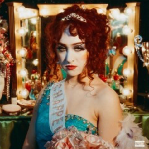 An album cover featuring a person with red hair and a blue dress, posing in front of a mirror with reflection showing trees. The person is wearing a sash and crown, suggesting a pageant theme.