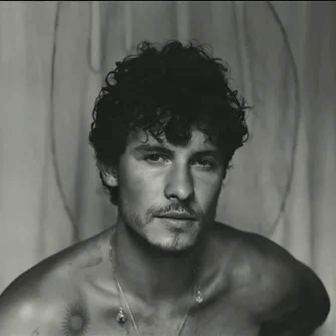 A black and white image of Shawn Mendes, shirtless, with a necklace and a subtle shadow behind him.