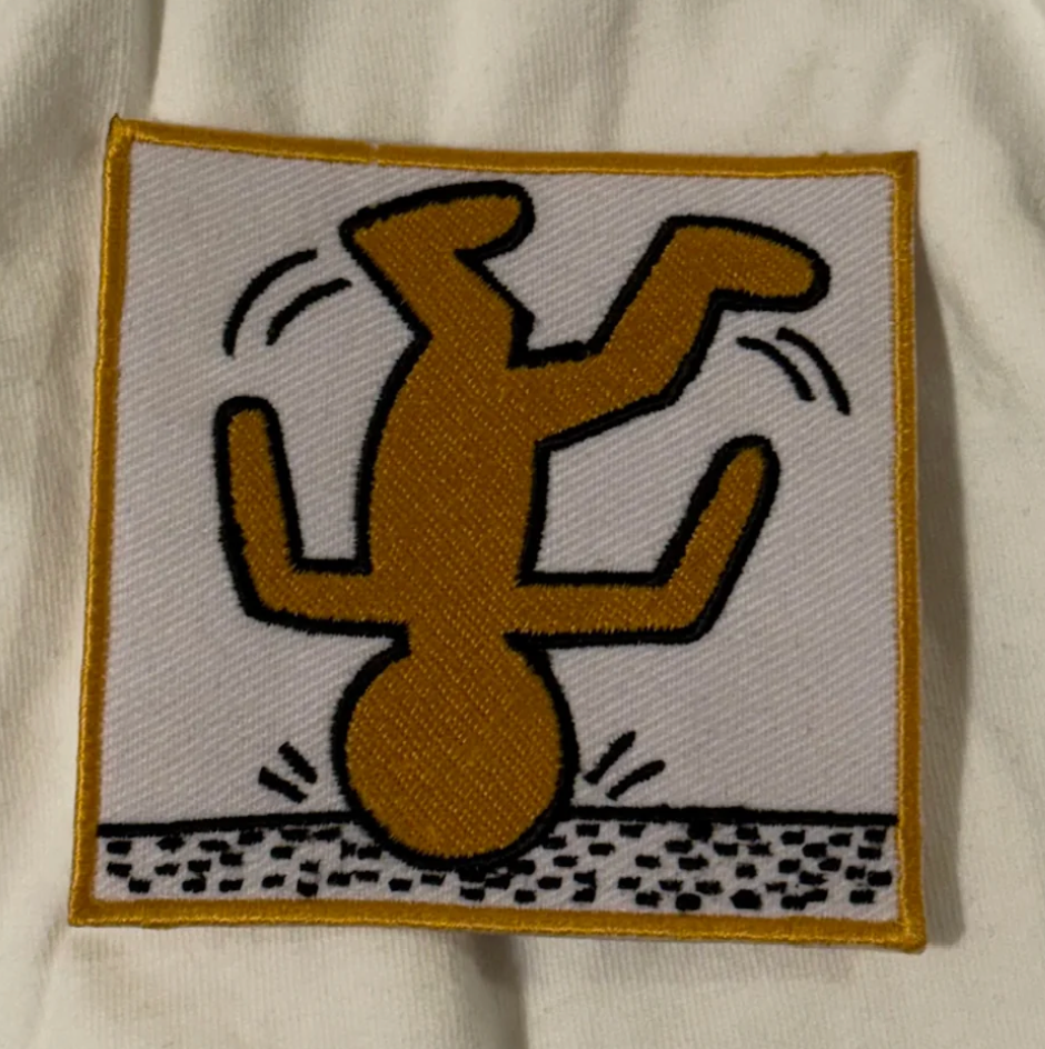 An embroidered square patch featuring a stylized upside-down figure in mustard yellow with black outlines on a white background, bordered in yellow, placed on a white fabric surface.