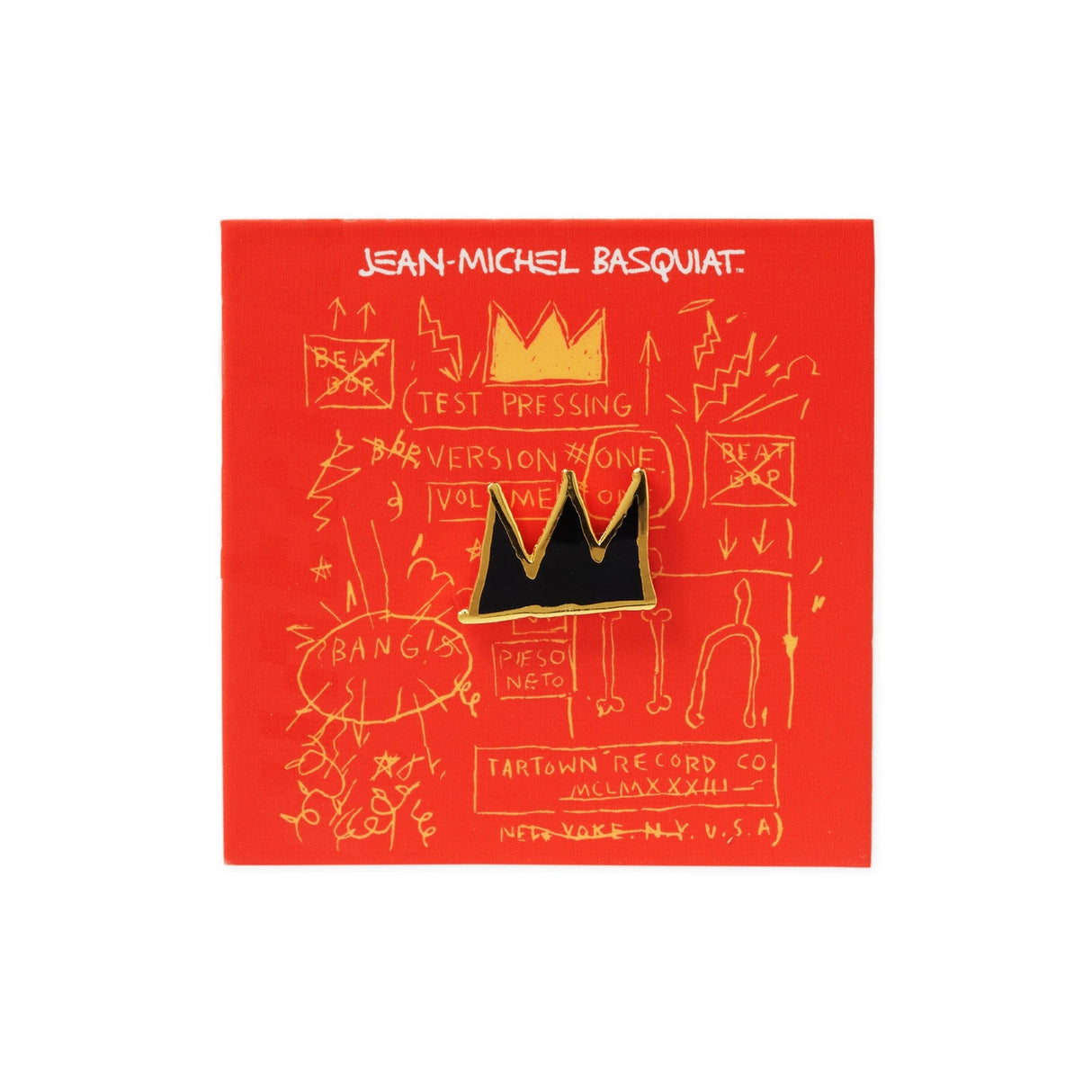A black and gold crown-shaped enamel pin by Jean-Michel Basquiat, mounted on a red backing card with gold graffiti-style drawings and text.