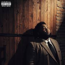 A man in a brown patterned blazer, white shirt, and black tie leans against a wooden door and brick wall, with a Parental Advisory label in the top-left corner.