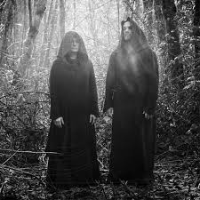 Two figures in dark cloaks standing in a forest with a sunbeam effect.