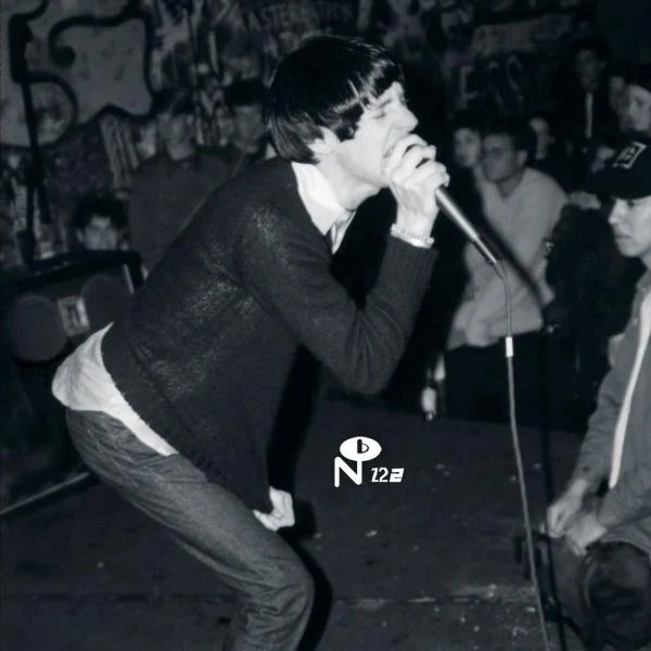 Black and white photo of a person singing into a microphone on a stage, wearing a dark textured sweater and jeans, with a crowd and graffiti in the background.