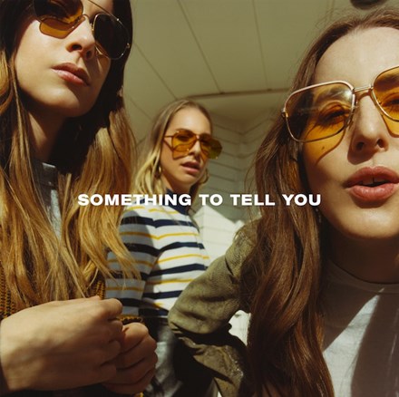The image features the cover art for "Something To Tell You," the sophomore album by Haim. The cover is styled with a close-up shot of the three band members, Danielle, Este, and Alana Haim, displaying a casual and confident vibe.