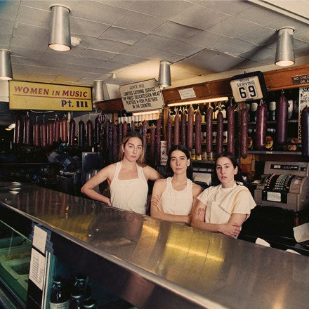 Three women standing behind a counter in a deli setting, wearing aprons, with a sign in the background indicating 'Women in Music Pt. 11'.