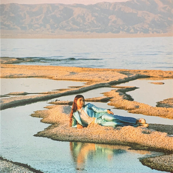 An album cover featuring a person sitting on a rocky surface with a calm lake and mountains in the background.