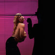 A woman in a black outfit poses behind horizontal blinds against a vivid pink background, while a large black silhouette of a person in a suit and hat stands on the right holding a thin object toward her face.