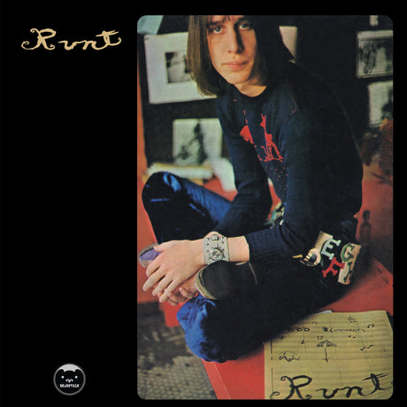 Runt - The Art Album cover, featuring a portrait of a young man with long brown hair sitting cross-legged on a red bench. He is wearing a dark blue sweater with a red graphic and blue jeans.