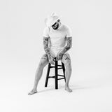 A black and white studio portrait of a man sitting on a stool, wearing a white cowboy hat, white t-shirt, and white shorts, with visible tattoos on his arms and legs.
