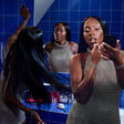 A woman with long black hair applies makeup while looking into a mirror in a blue-tiled bathroom. She wears a shimmering metallic taupe dress, and her reflection is visible in the mirror.