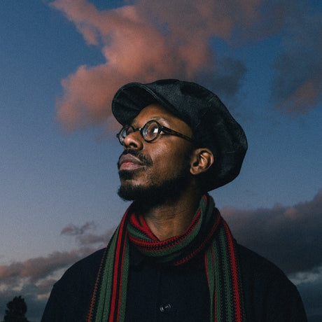 Man wearing a black cap and colorful scarf against a dramatic sky with clouds.