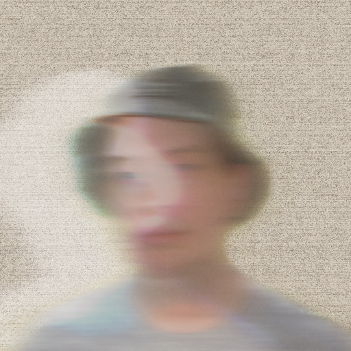 Blurred image of a person wearing a cap against a textured beige wall.