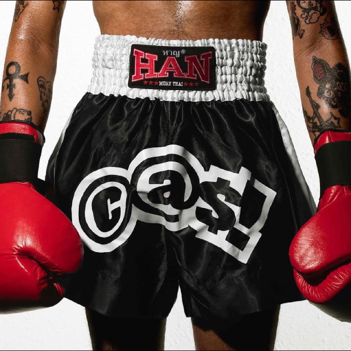 Person wearing black boxing shorts with white logos and red boxing gloves, on a plain background