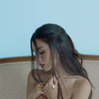 Woman with long hair looking down, wearing a gold necklace against a light blue background