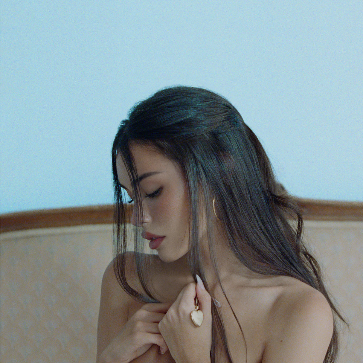 Woman with long hair looking down, wearing a gold necklace against a light blue background
