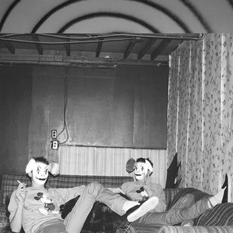 Two people wearing masks sitting on a couch in a room with curtains and a light switch.