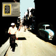 Album cover art for Buena Vista Social Club's 20th Anniversary World Tour, featuring a man walking down a sunlit street with vintage cars on the right, with a vinyl record sleeve graphic overlaid in the top-left corner.