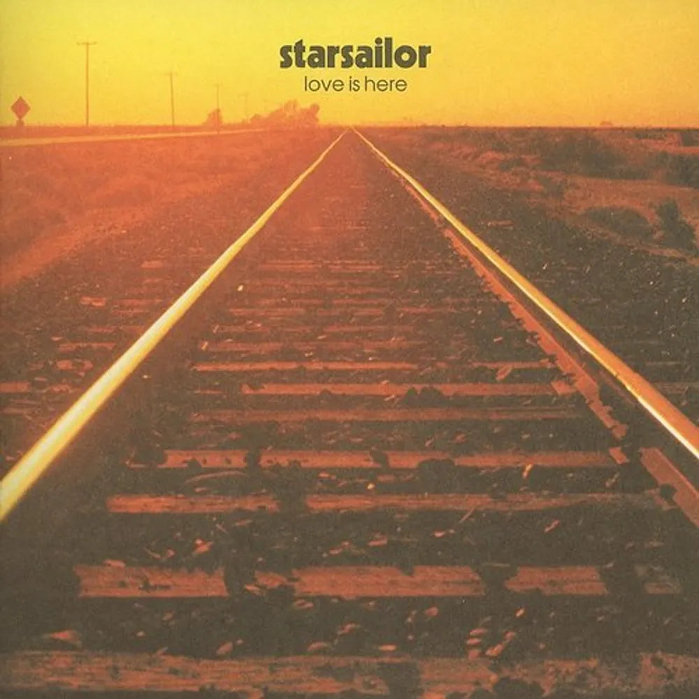 Album cover for 'love is here' by starsailor, featuring a photograph of railroad tracks receding into a desert landscape under a warm yellow-orange sky.
