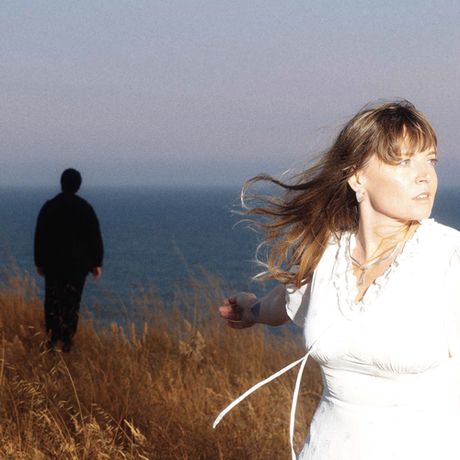 Woman in a white dress standing on a grassy hill with a blurred figure in the background.