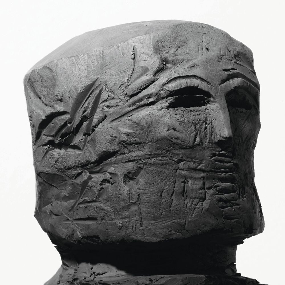 A dark charcoal-colored sculptural bust of a face with highly textured, weathered details, set against a plain white background.