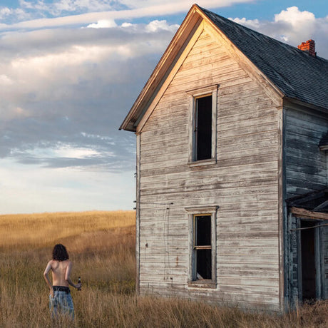Person standing in a field near an old wooden house with a cloudy sky.