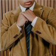 Man adjusting his tie wearing a brown corduroy jacket against a wooden panel background