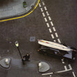 An overhead street scene showing a black car carrying a surfboard on its roof, with a person standing near a lamp post on the road. White dashed lane markings, a sidewalk, and a curb with yellow line are visible.