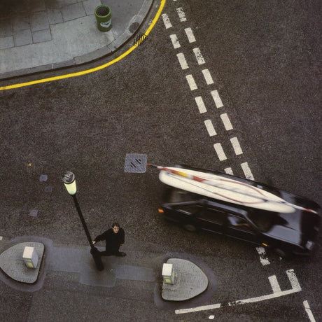 An overhead street scene showing a black car carrying a surfboard on its roof, with a person standing near a lamp post on the road. White dashed lane markings, a sidewalk, and a curb with yellow line are visible.