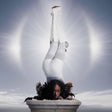 Person in a white outfit performing a handstand on a pedestal against a light gray background