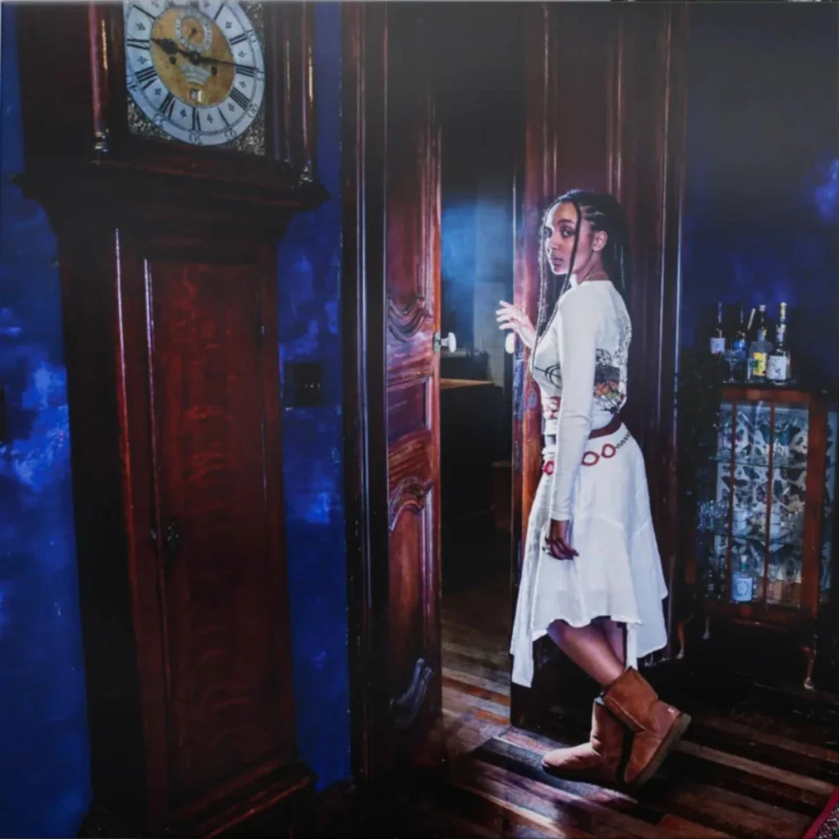 A woman in a white dress and brown boots stands in a doorway inside a dark room with blue lighting, next to a large wooden grandfather clock and a cabinet with bottles.