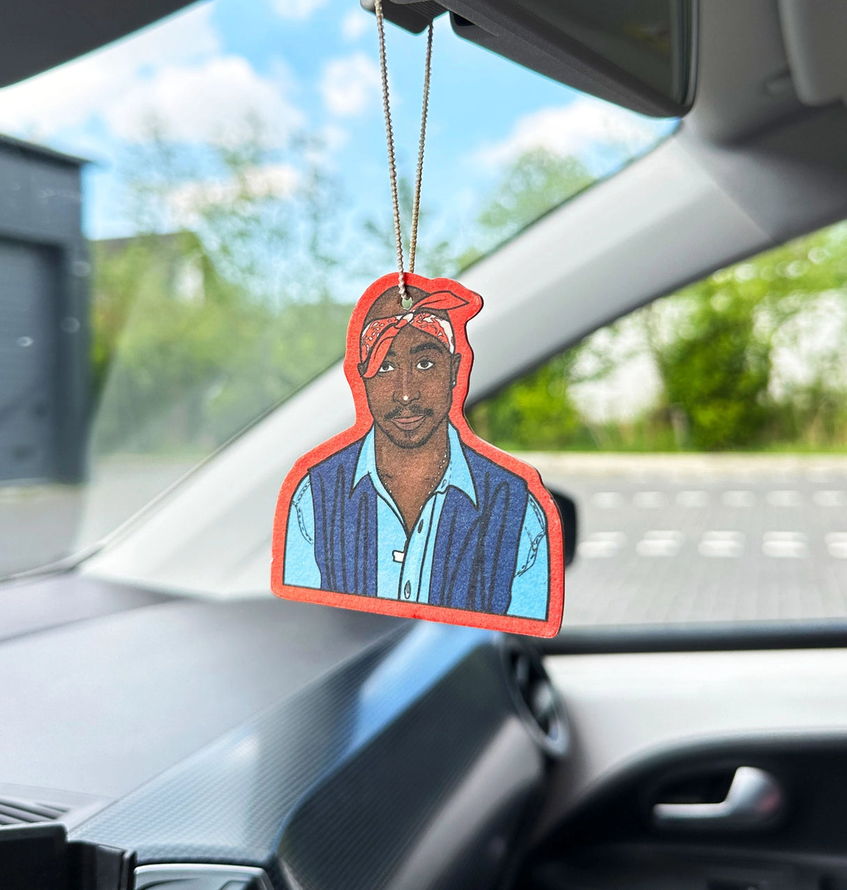 A die-cut air freshener or car fragrance hanging from the rearview mirror inside a car. The product features a stylized portrait of Tupac Shakur with a red outline, blue shirt, and dark vest, set against a blurred car interior and outdoor background.