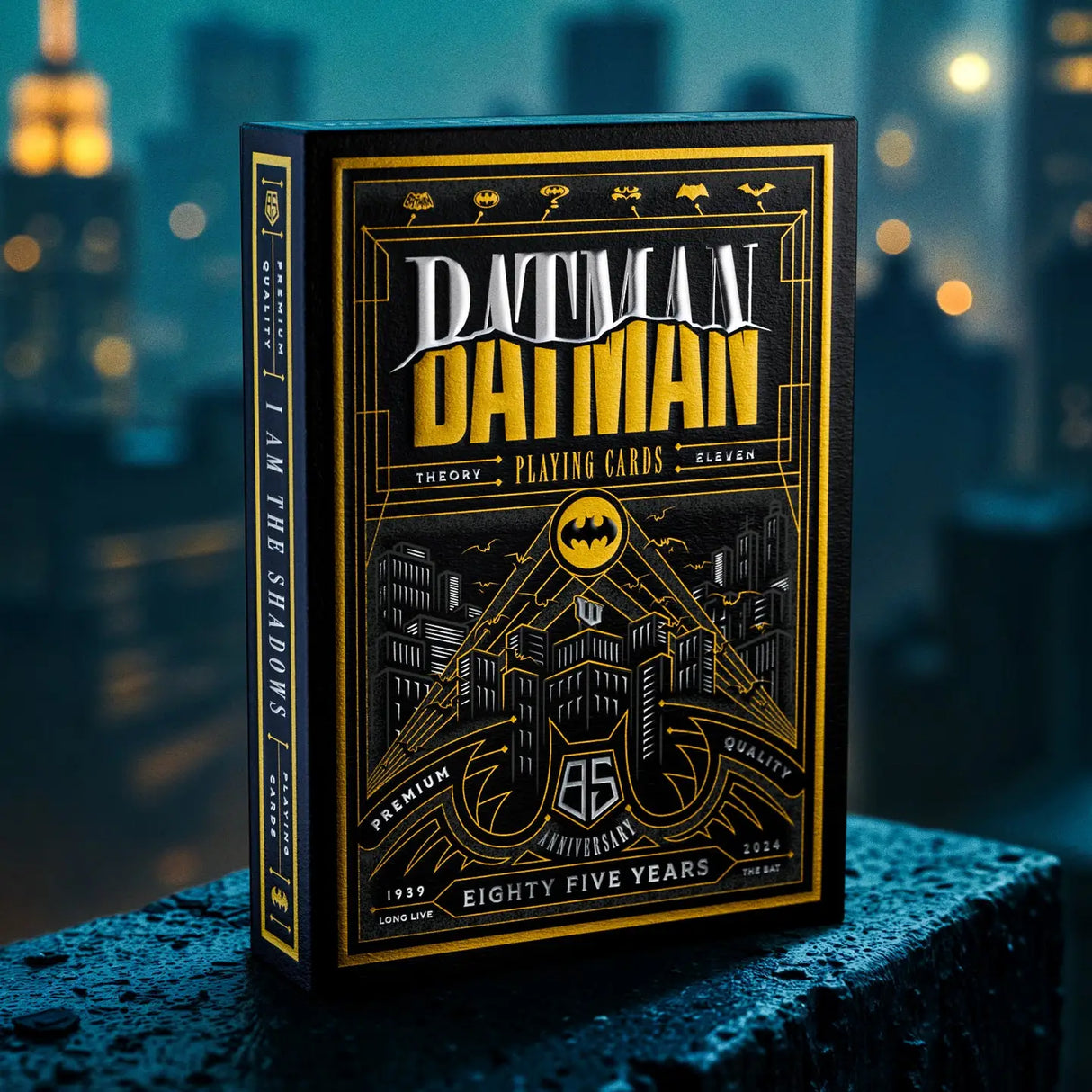 A boxed deck of Batman-themed playing cards stands upright on a wet dark surface with a blurred city skyline at night in the background. The box is black with prominent gold and white artwork and text reading 'Batman' and 'The Theory Playing Cards'.