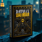 A boxed deck of Batman-themed playing cards stands upright on a wet dark surface with a blurred city skyline at night in the background. The box is black with prominent gold and white artwork and text reading 'Batman' and 'The Theory Playing Cards'.