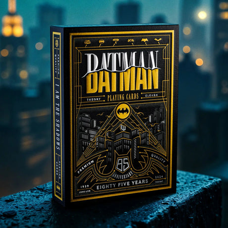 A boxed deck of Batman-themed playing cards stands upright on a wet dark surface with a blurred city skyline at night in the background. The box is black with prominent gold and white artwork and text reading 'Batman' and 'The Theory Playing Cards'.