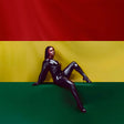 Person in a black latex outfit sitting against a red, yellow, and green striped background